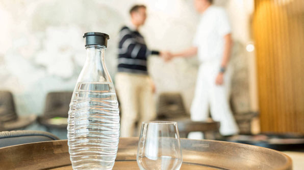 Wasserflasche mit Glas im Vordergrund und Arzt begrüßet Patienten im Hintergrund